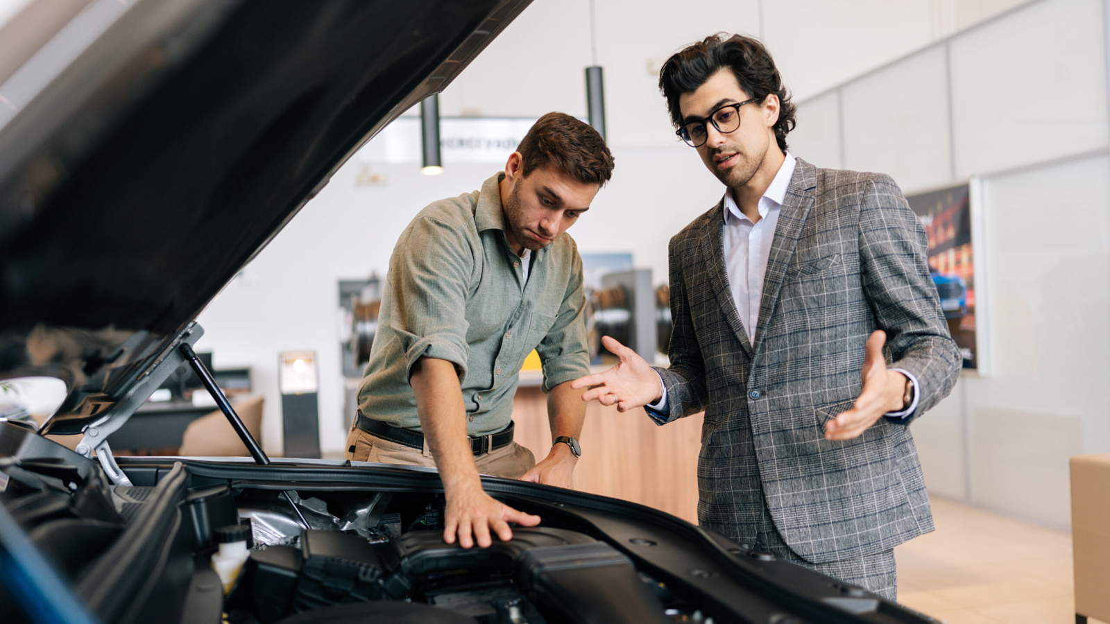 Portrait of professional dealer manager in business suit and male client near new car with open hood. Stylish friendly dealer talking about technical characteristics. Concept of choosing new auto
